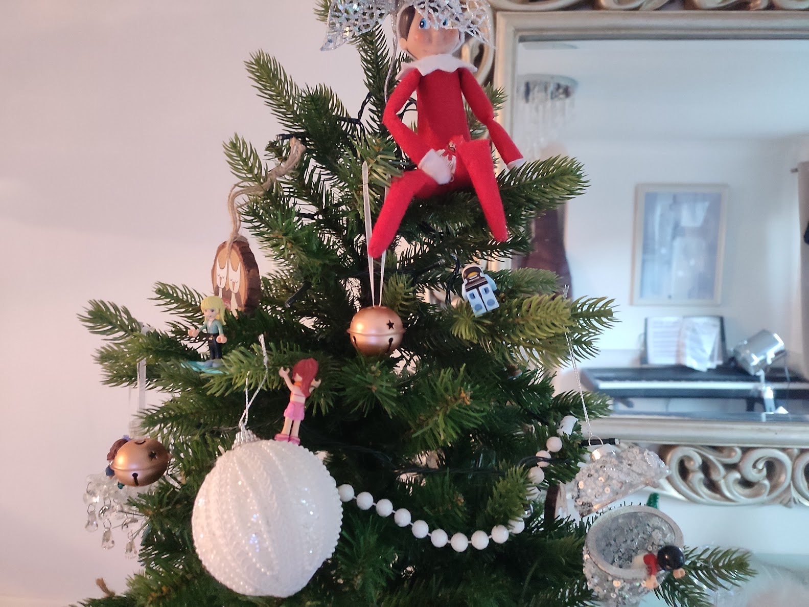 A Christmas elf sits at the top of a decorated tree while small lego figures climb the branches below, forming a funny festive scene.
