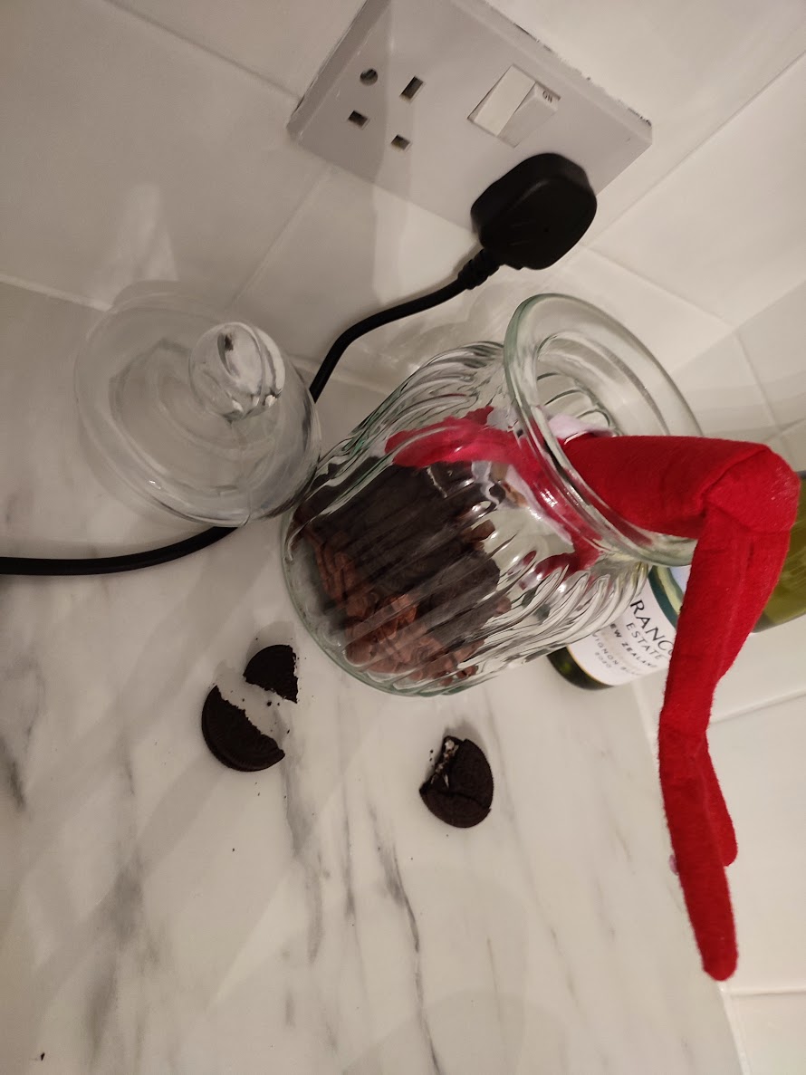 A Christmas elf is upside down in a cookie jar with broken biscuits and crumbs scattered around, caught mid-cookie raid in a funny festive scene.