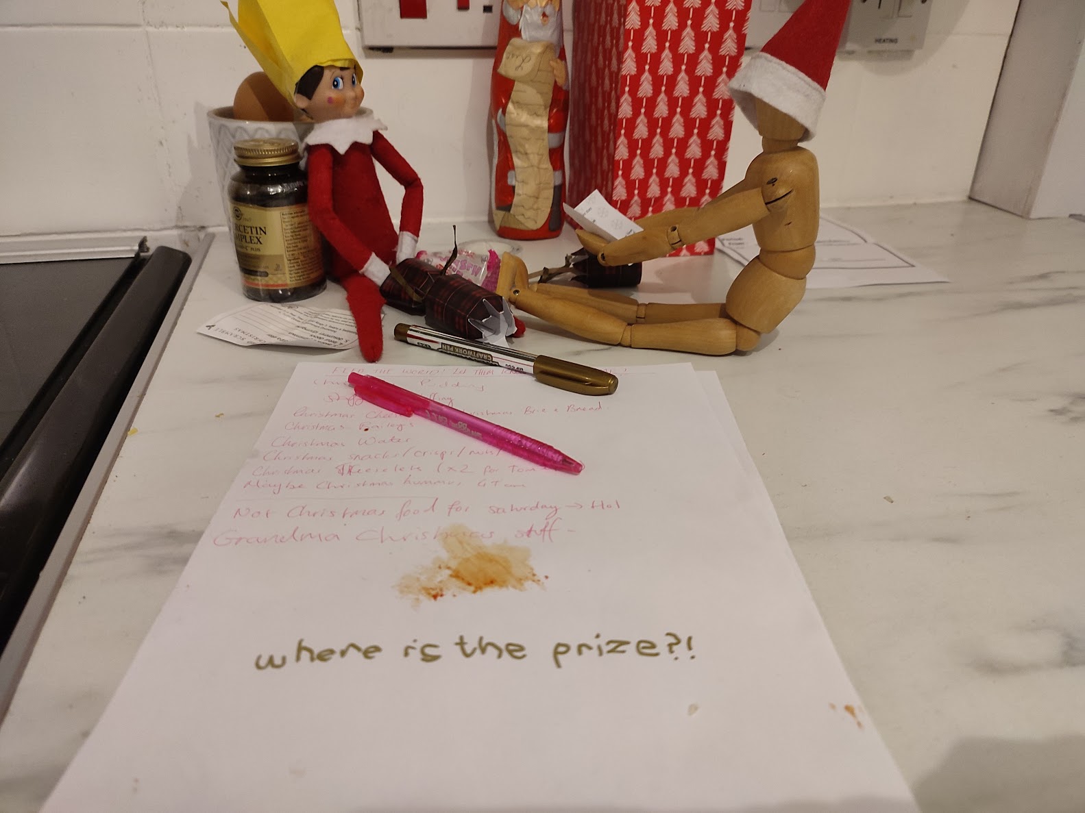 A Christmas elf wearing a cracker hat sits beside a toy wearing the elf’s hat, with a pulled cracker and a joke between them in a funny festive setup.