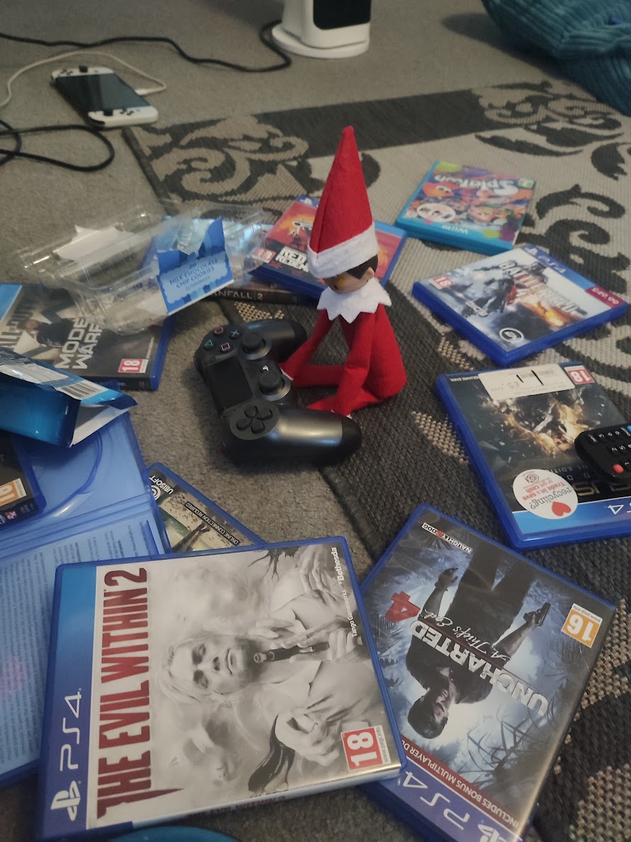 A Christmas elf sits propped against a games console remote surrounded by video games, with the tv screen glowing as if he’s been gaming all night.