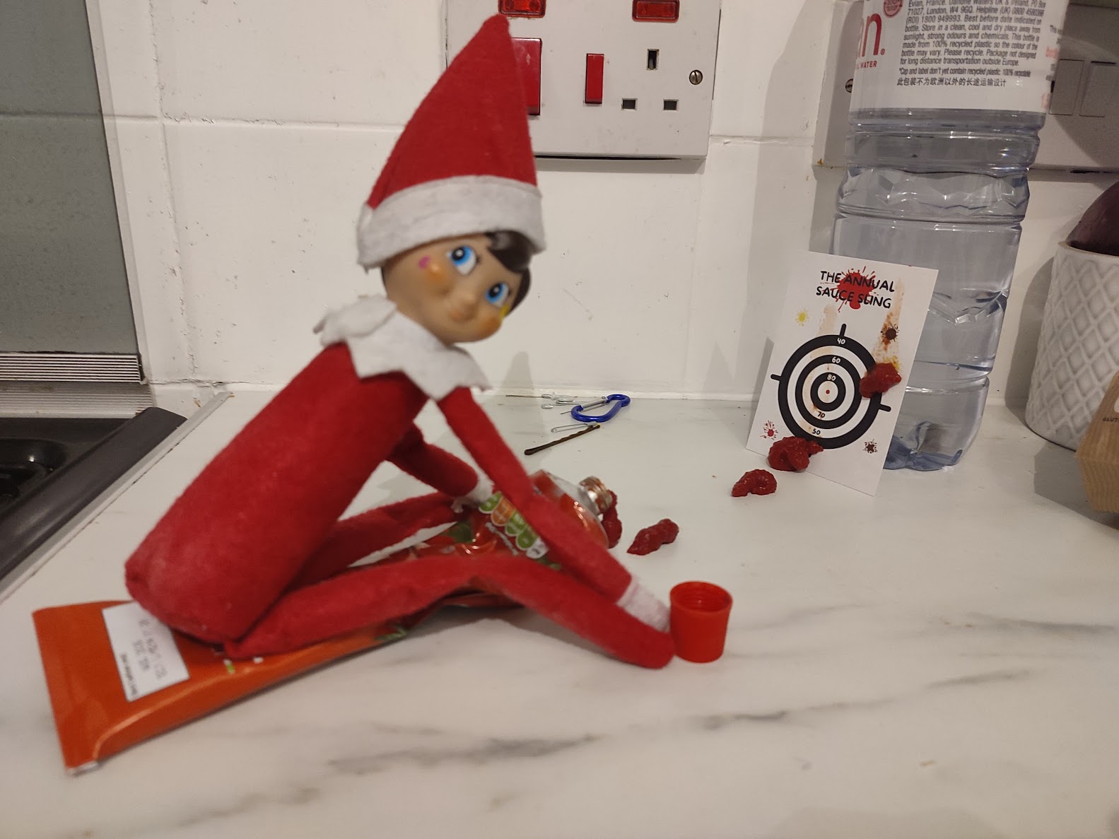 A naughty Christmas elf sits on a ketchup bottle pointed at a target card labelled “The Annual Sauce Fling,” with splashes of sauce on the target.