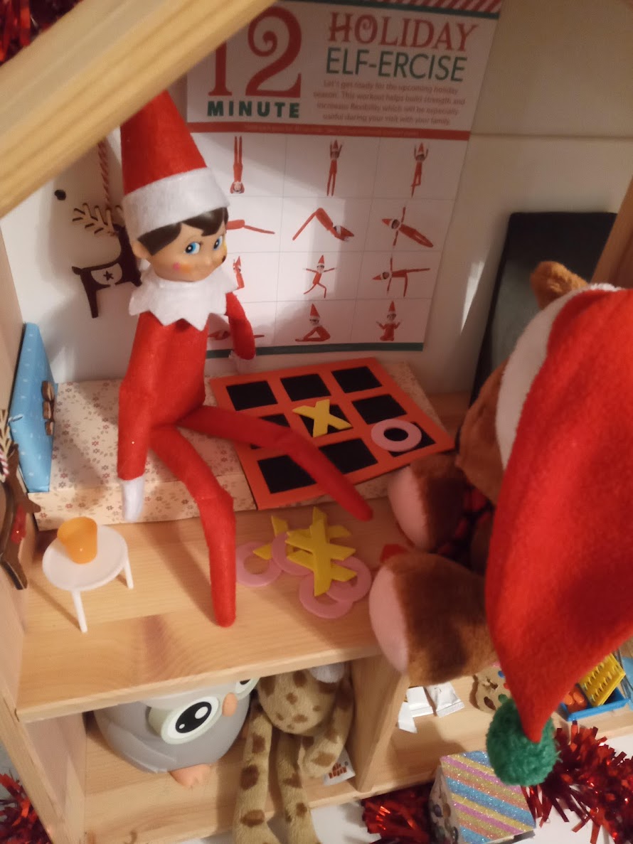 A Christmas elf sits with a soft toy friend playing noughts and crosses using a small board with Xs and Os between them.