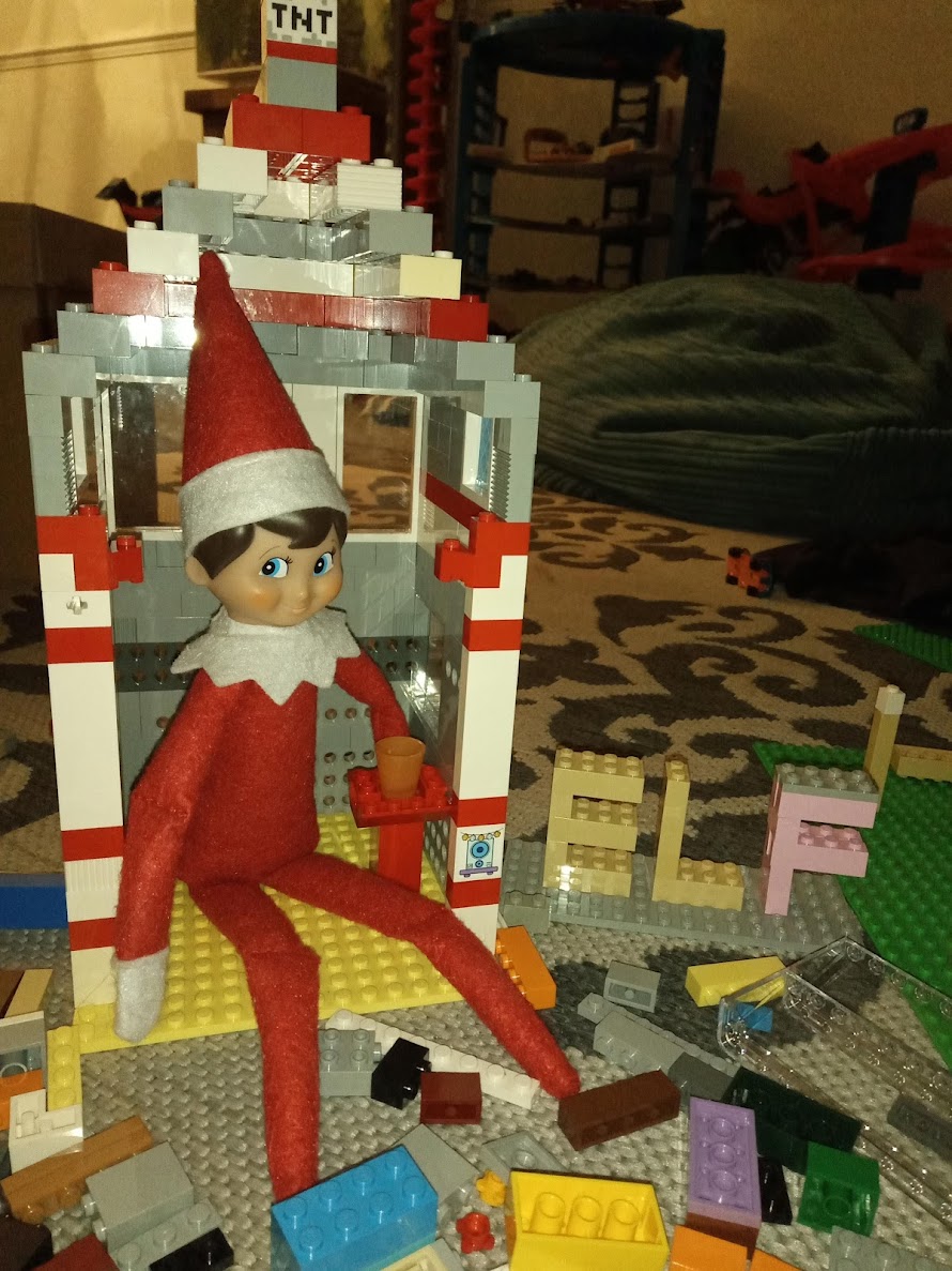 A Christmas elf sits beside a Lego house with a pointy roof for his hat, a TNT block on top and the word “ELF” spelled out in Lego bricks.