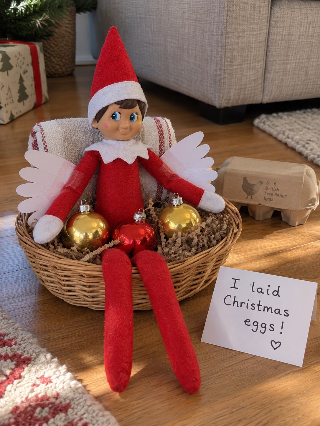Tom’s elf with white paper wings seated in a wicker nest beside three Christmas baubles and an I laid Christmas eggs note.