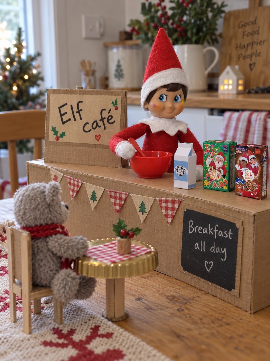 Tom’s elf behind a cardboard breakfast café counter with miniature cereal boxes, milk carton and red bowl, serving a teddy customer.
