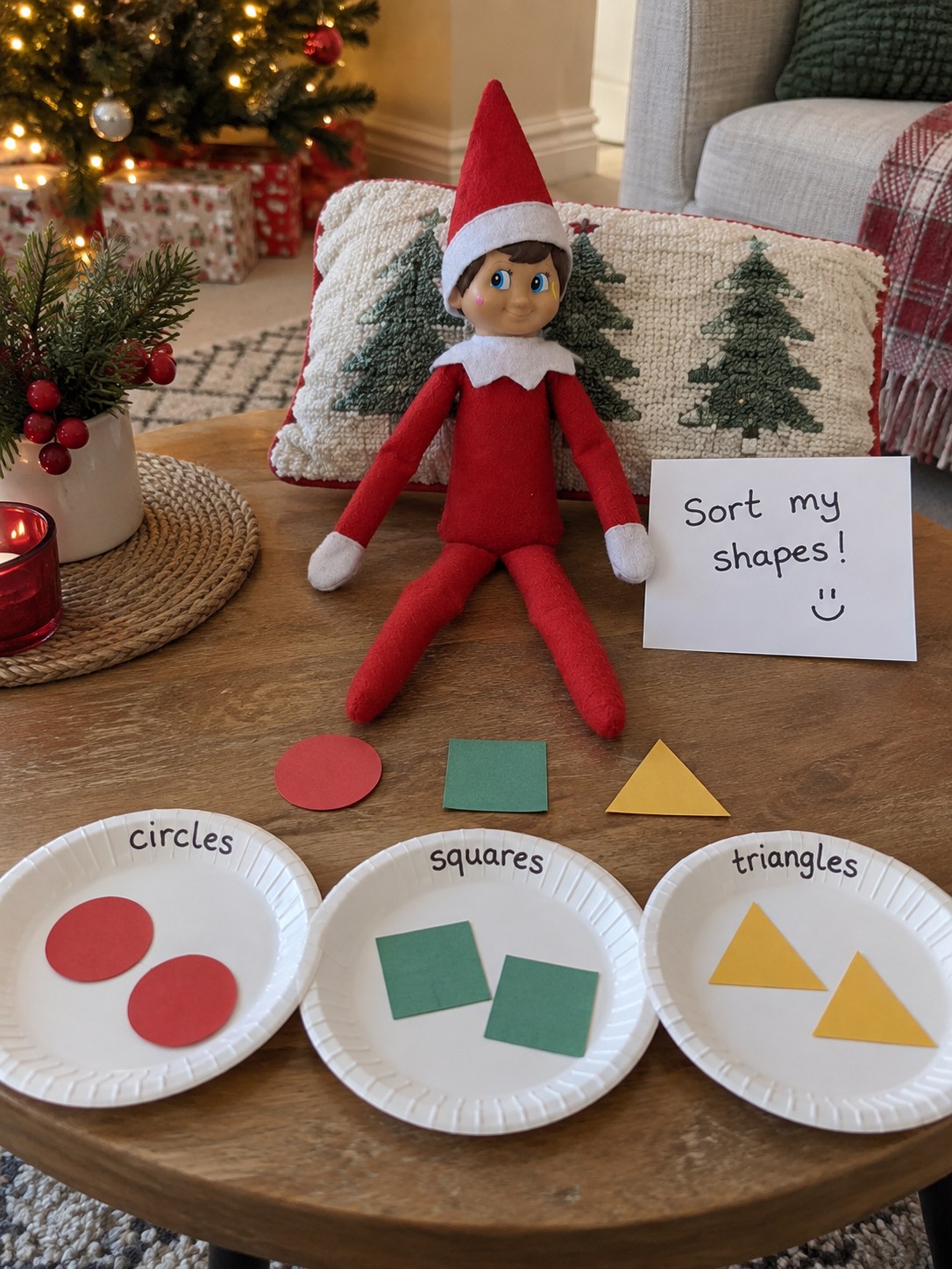 Tom’s elf behind three paper plates labelled circles, squares and triangles, holding red circles, green squares and yellow paper triangles.
