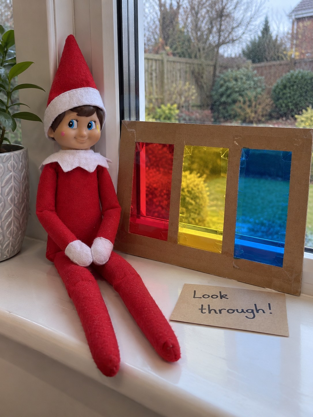 Tom's elf on a windowsill beside a homemade card frame with three transparent red, yellow and blue panes.