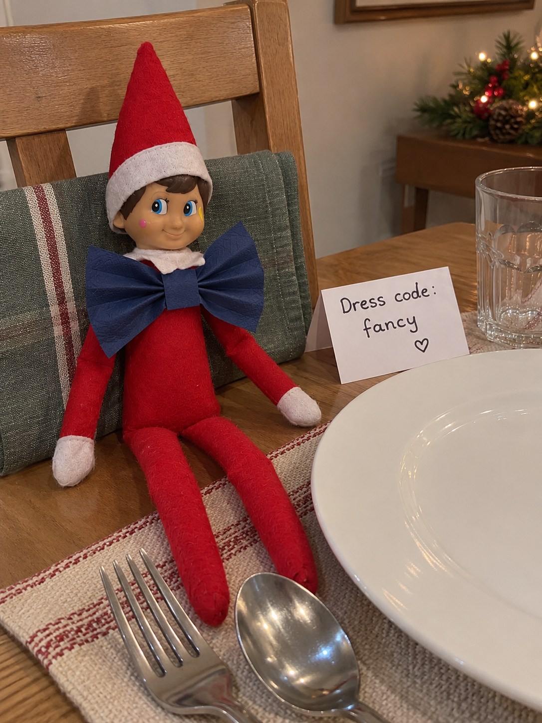 Tom's elf wearing a large dark-blue paper-napkin bow tie beside an empty Christmas dinner place setting.
