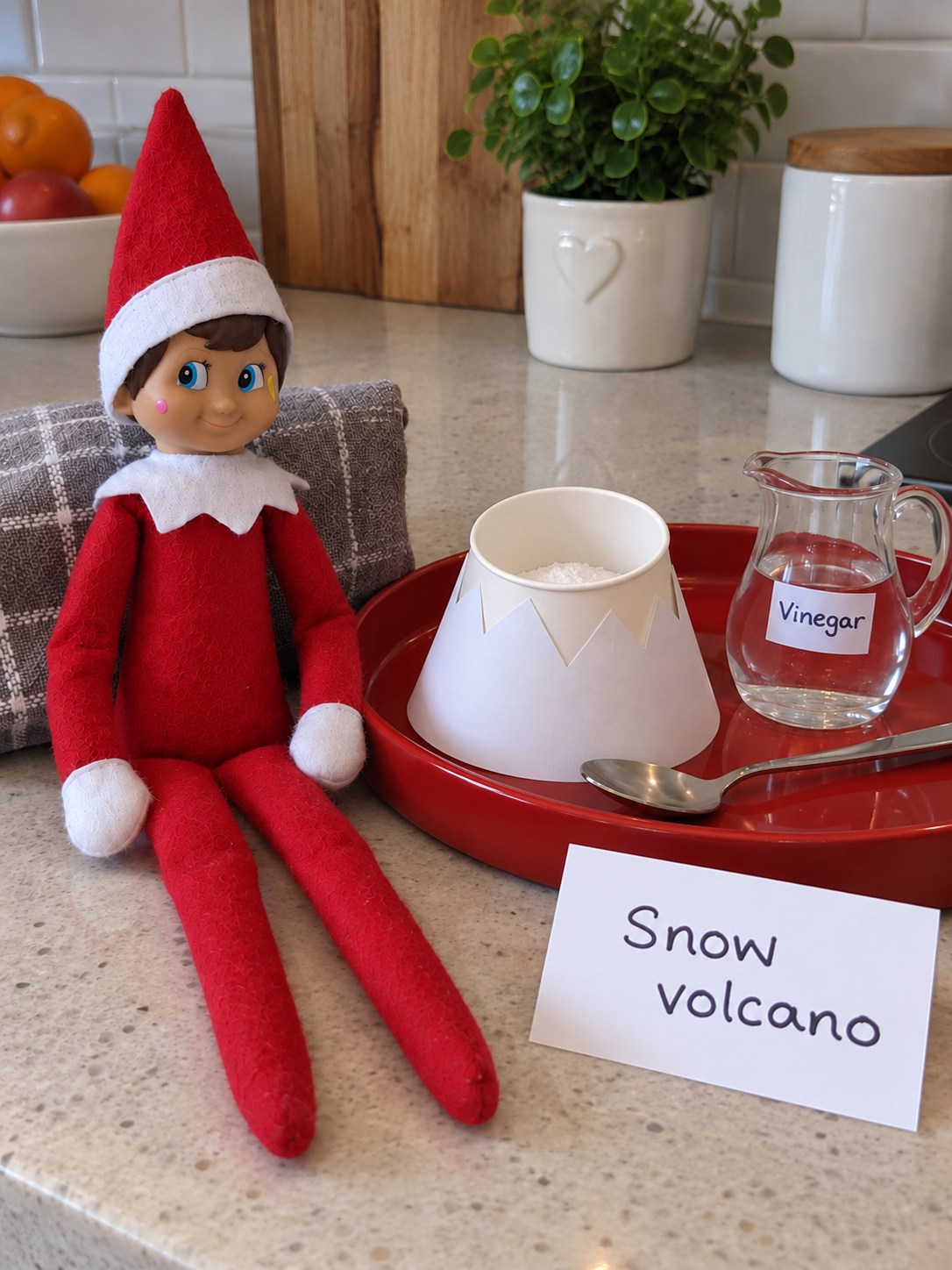 Tom’s elf beside an open paper cup inside a white snow-mountain sleeve, a vinegar jug, a teaspoon and a Snow volcano note on a red tray.