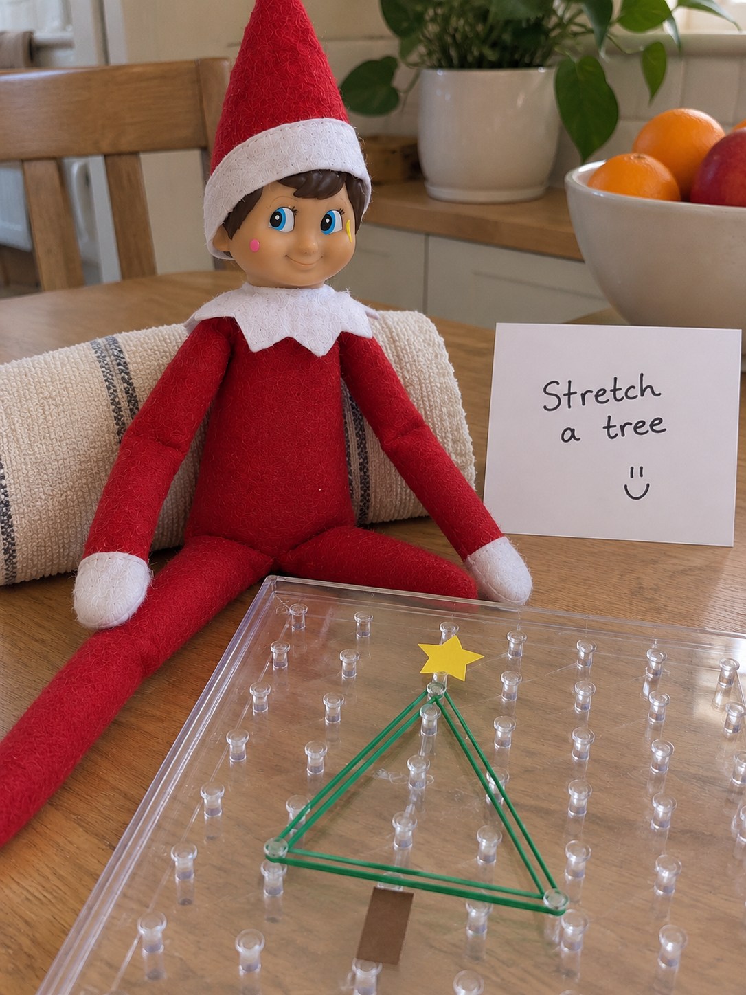 Tom’s elf beside a clear peg geoboard with green elastic bands outlining a triangle, a paper trunk and a Stretch a tree note.
