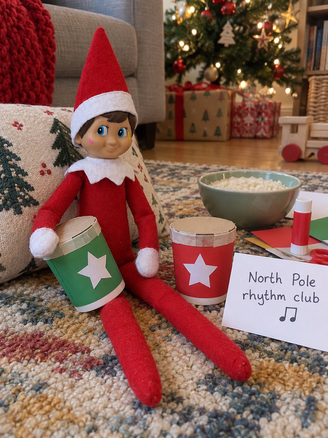 Tom’s elf on a rug beside two sealed yoghurt-pot shakers decorated with red and green paper sleeves and white stars.