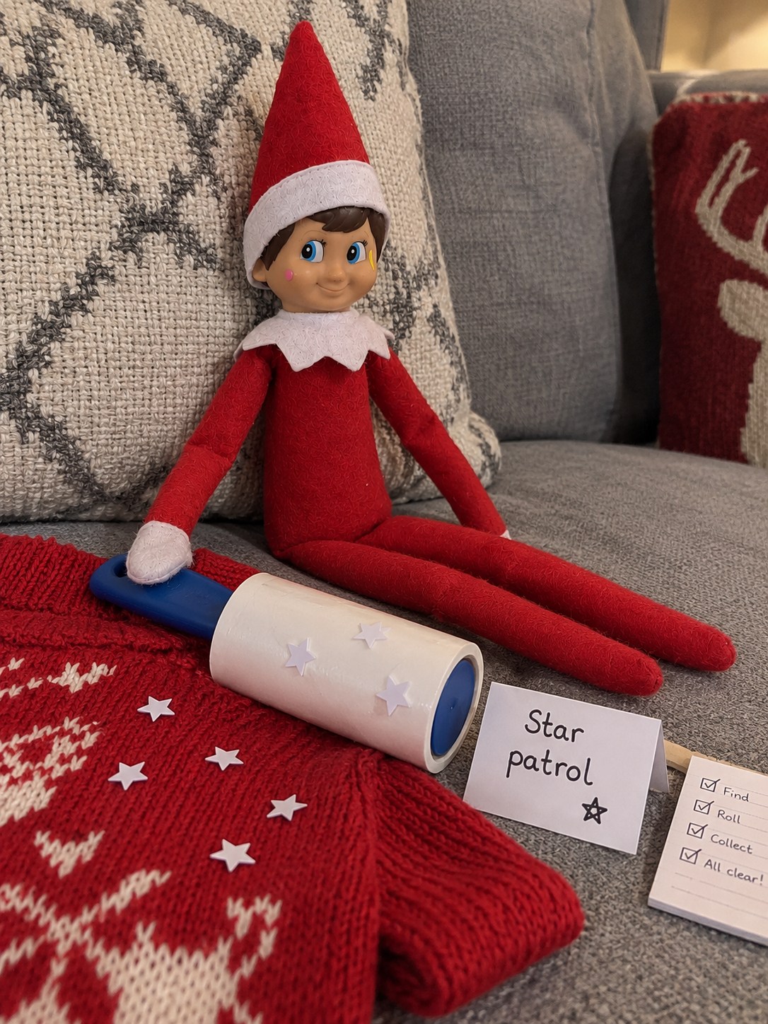 Tom’s elf beside a red Christmas jumper, a blue-handled lint roller and a few white paper star confetti pieces.