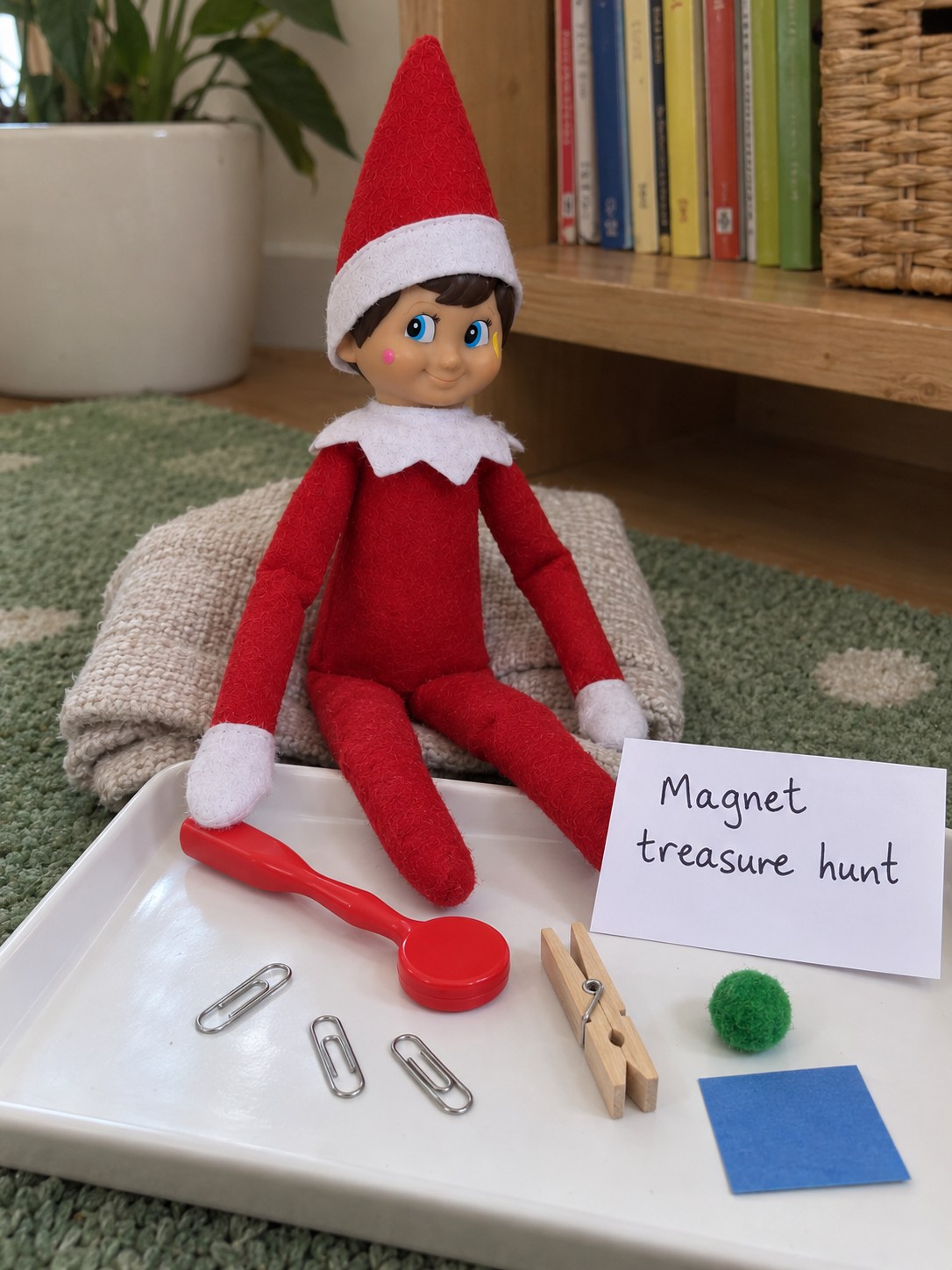 Tom’s elf beside a red round-headed magnetic wand, steel paper clips, a wooden peg, a green pom-pom and blue card on a white tray.