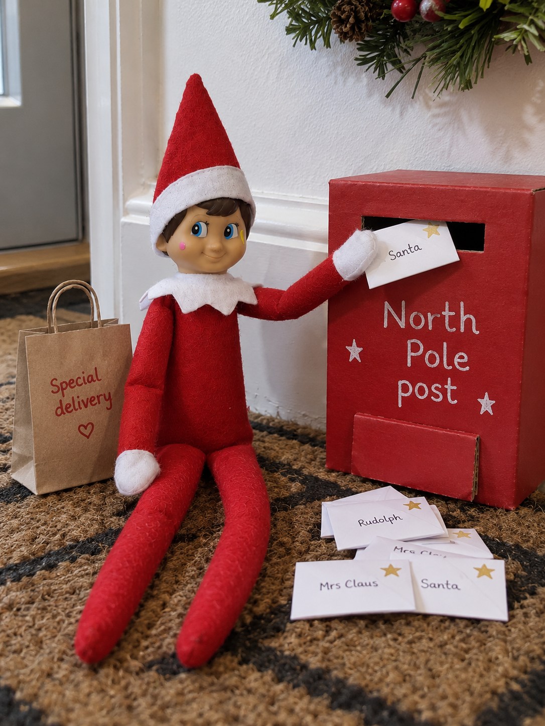 Tom’s elf beside a homemade red North Pole postbox on a hallway mat, posting a tiny envelope with more letters nearby.