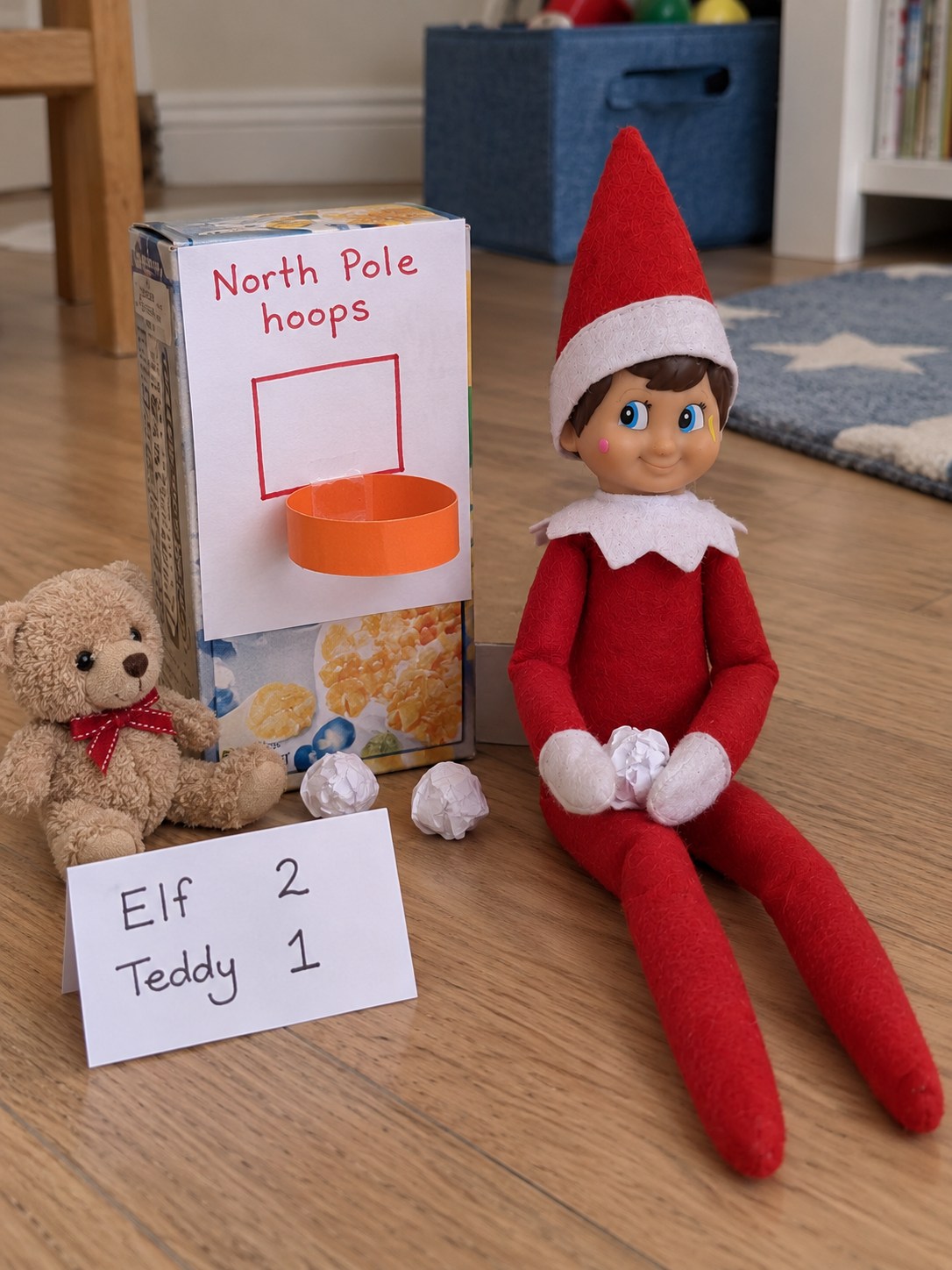 Tom’s elf holding a crumpled paper ball beside a homemade basketball hoop and backboard with a toy bear watching.