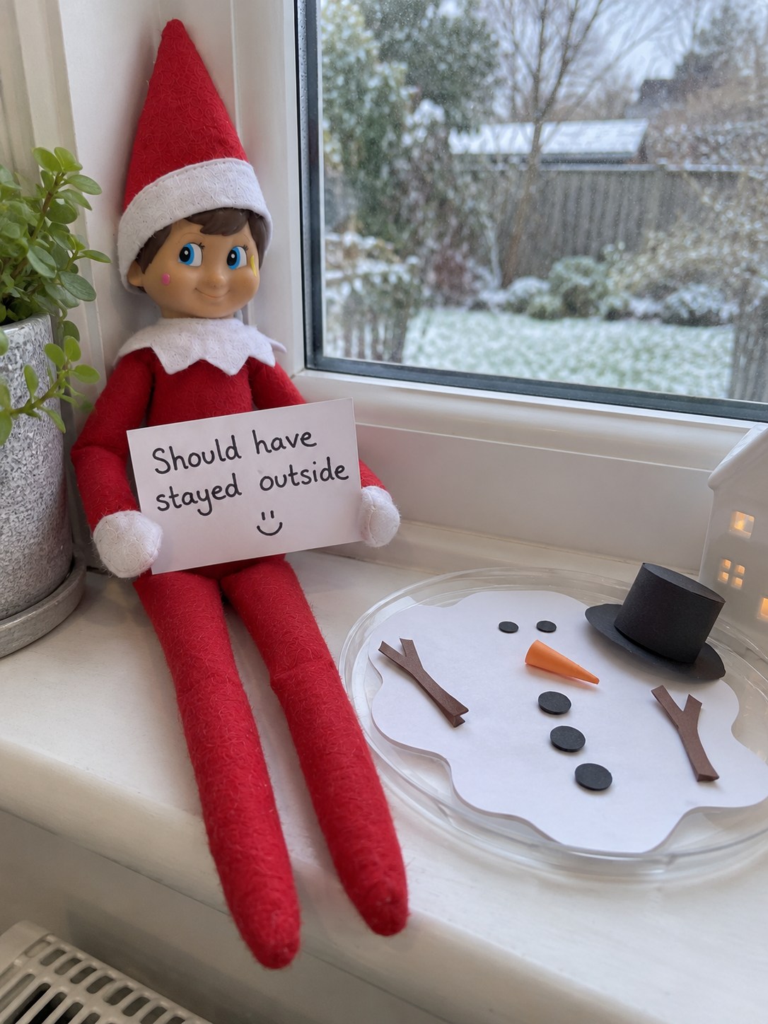Tom’s elf beside a flat white paper snowman puddle with black paper buttons, orange paper carrot nose, twig arms and a top hat.