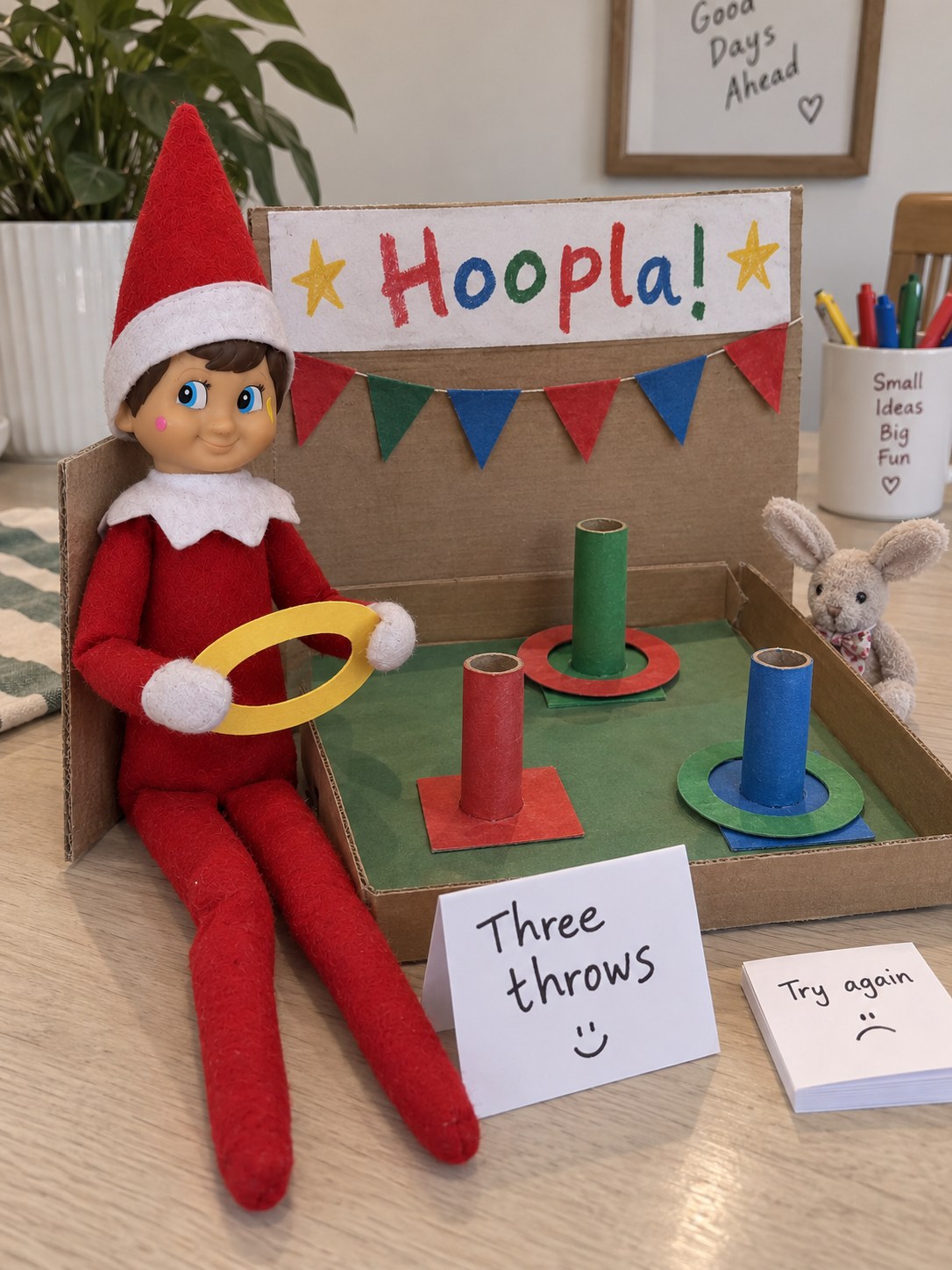 Tom’s elf holding a yellow paper ring beside a homemade hoopla game with three cardboard posts and red and green rings.