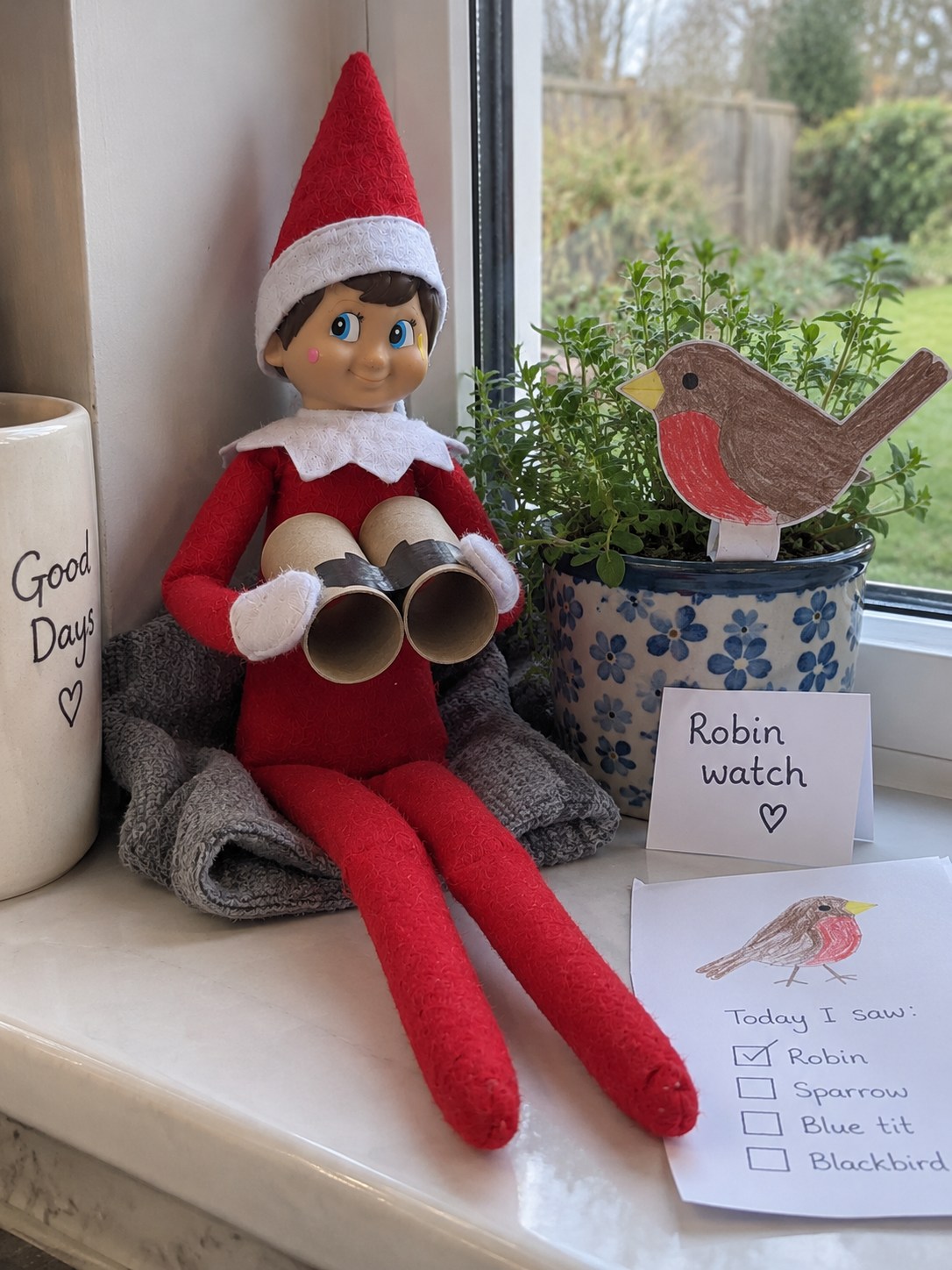 Tom’s elf holding two short cardboard-tube binoculars beside a flat paper robin on a plant pot and a Robin watch note.