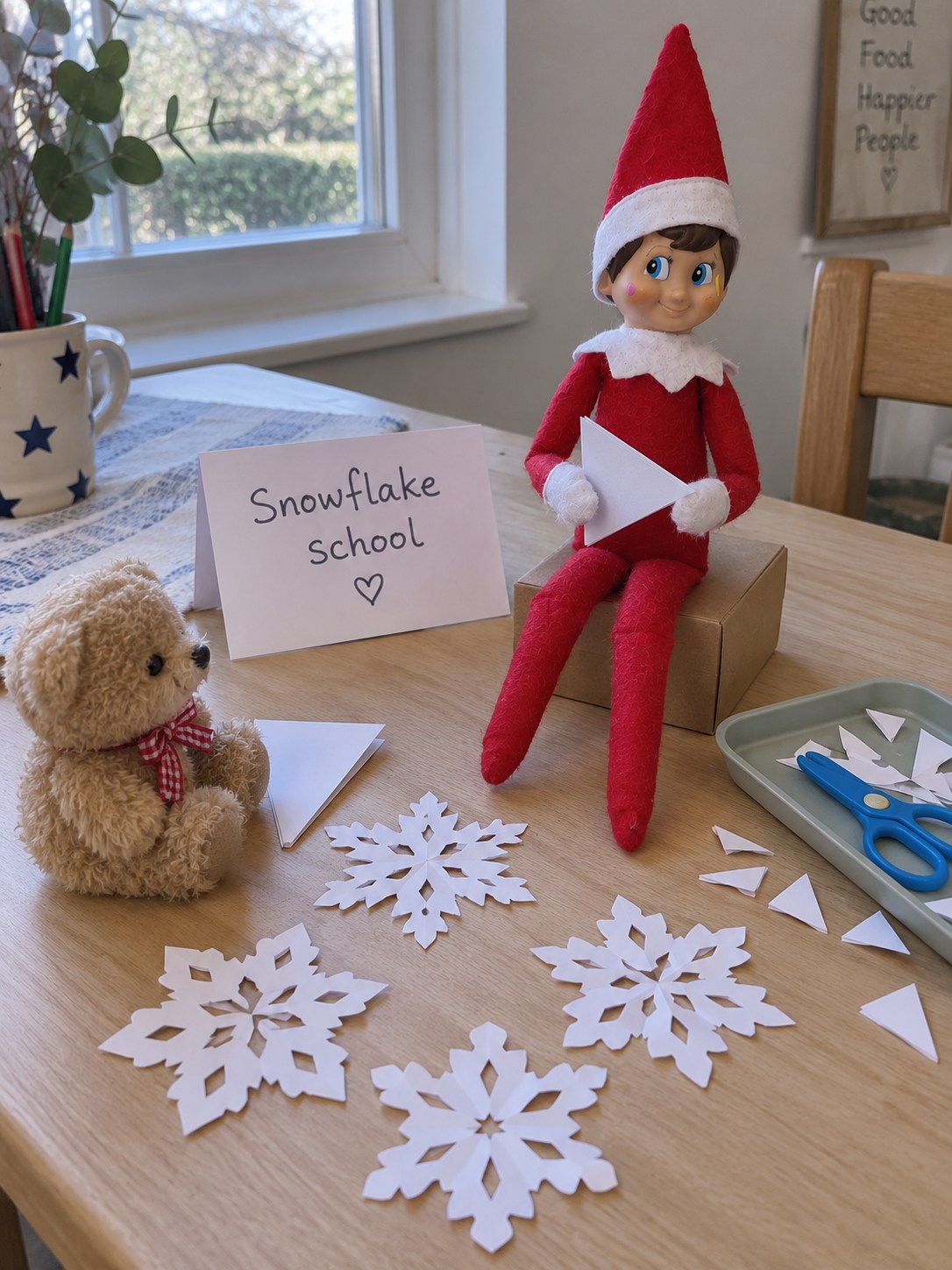 Tom’s elf holding folded white paper at a snowflake craft class with four cut-paper snowflakes, closed scissors and a teddy pupil.