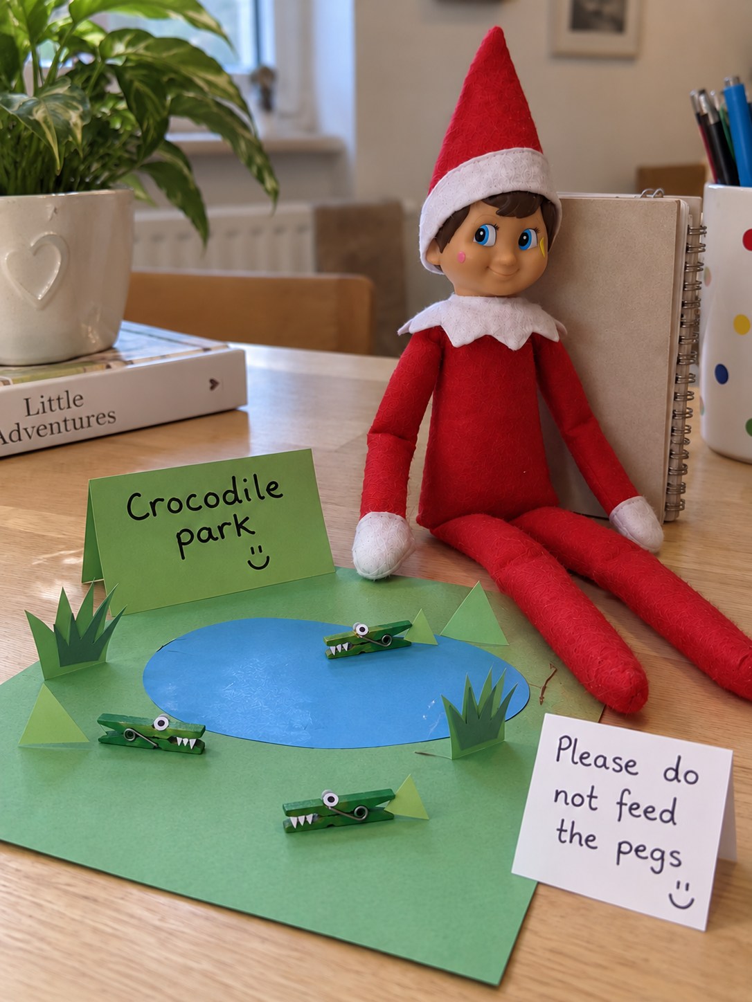 Tom’s elf beside three tiny green wooden peg crocodiles on a card park with a blue paper pond.