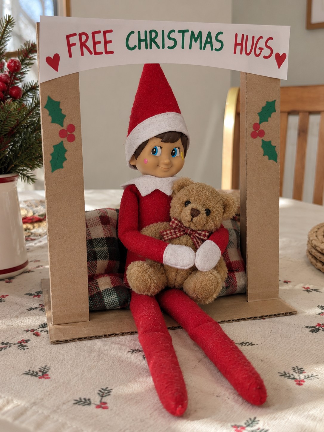 Tom's elf hugging a brown teddy inside a cardboard booth with a Free Christmas Hugs banner.