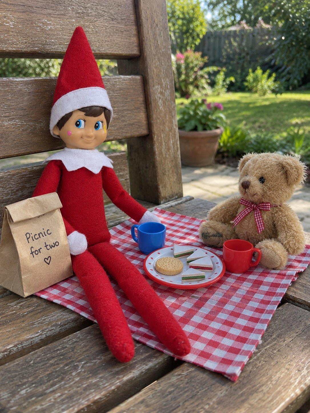 Tom’s elf and a teddy on a red-and-white handkerchief picnic blanket with paper sandwiches, a biscuit and two miniature cups.