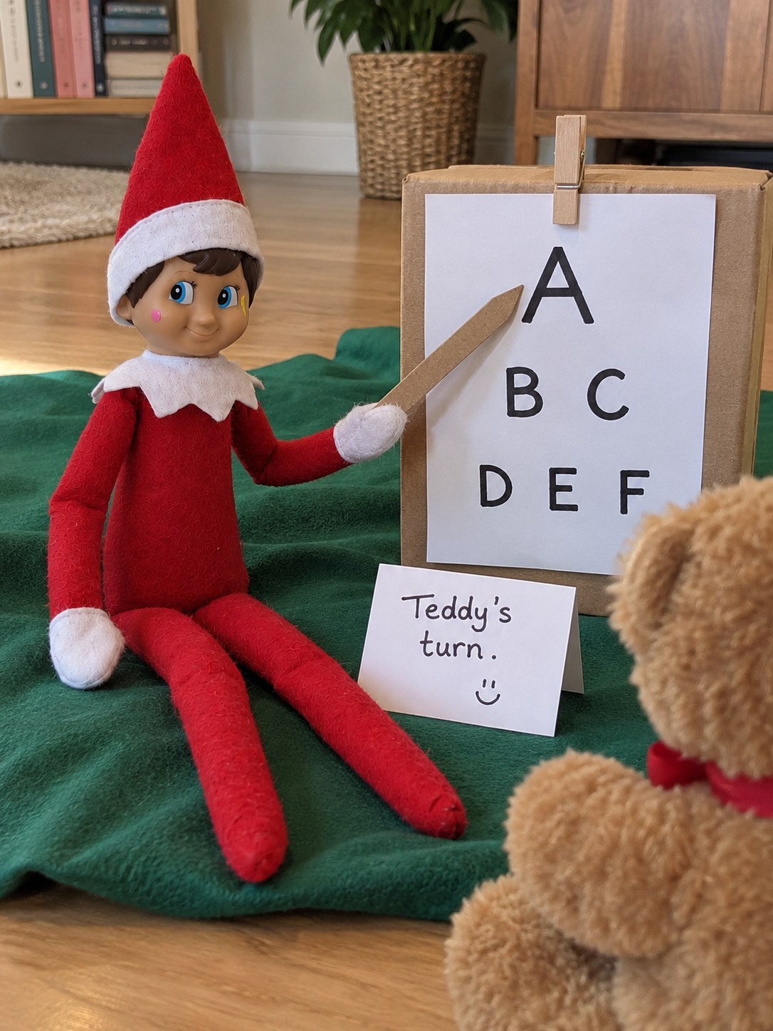 Tom’s elf pointing towards a homemade A, B C, D E F letter chart while Teddy sits opposite beside a Teddy’s turn note.