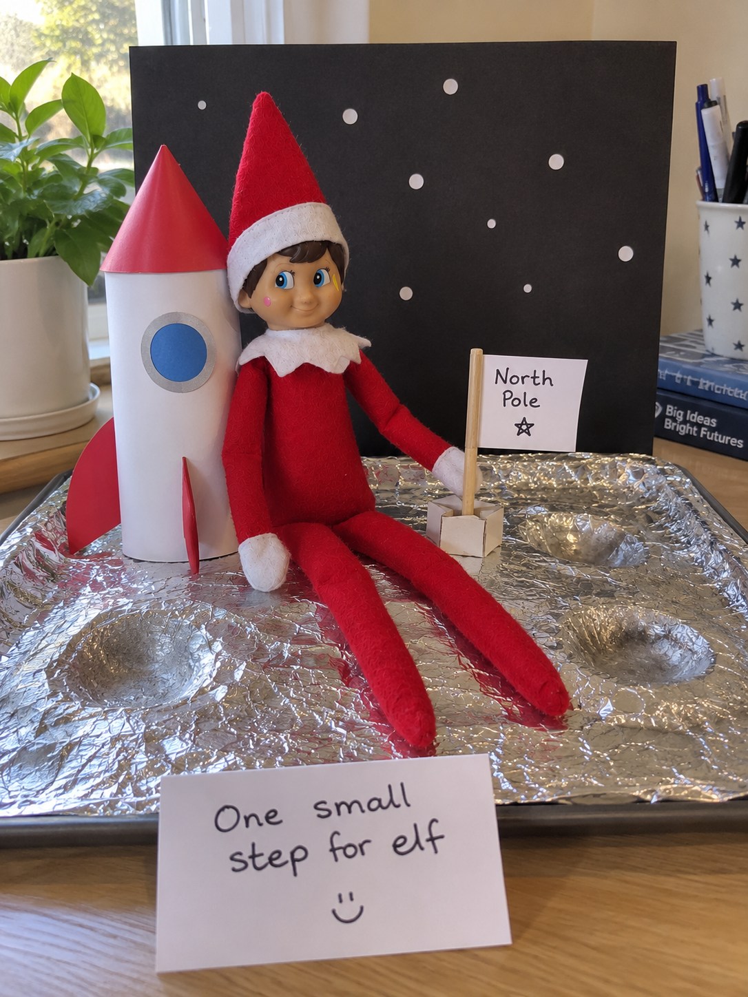 Tom’s elf seated on a crumpled-foil moon surface beside a homemade paper rocket and small North Pole flag.