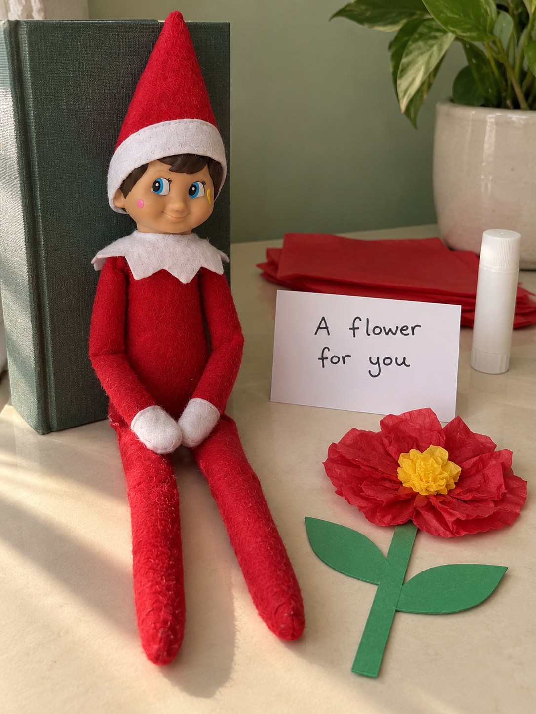 Tom's elf beside a homemade red tissue-paper flower with a yellow centre, green card stem and two leaves.