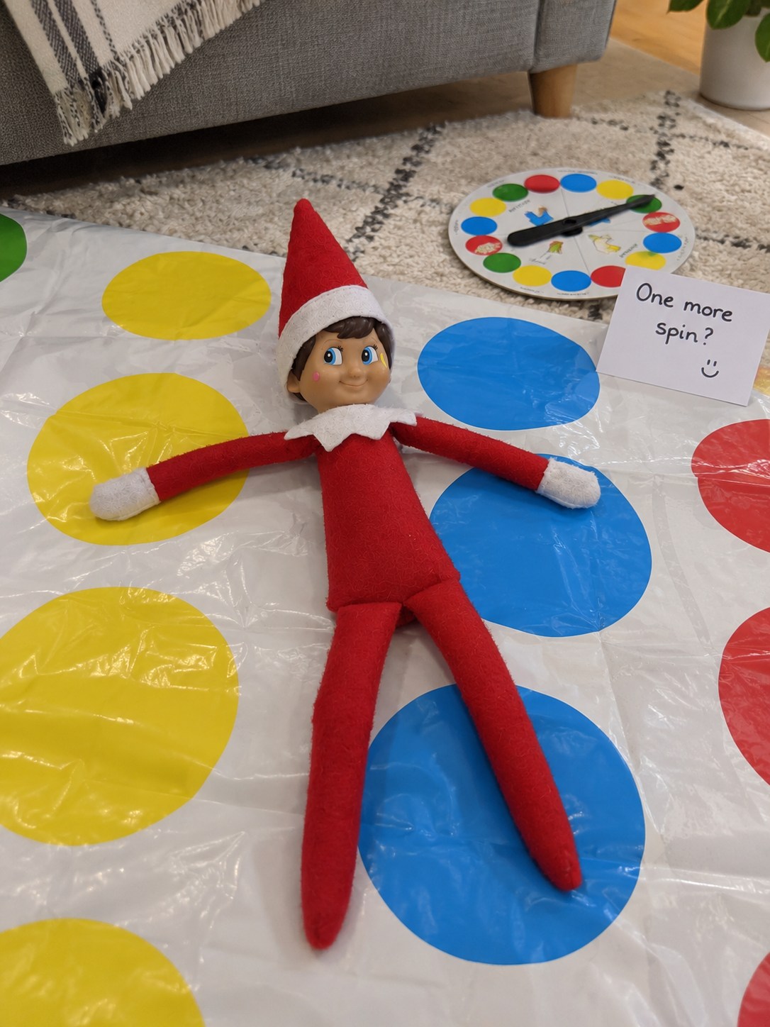 Tom’s elf lying on a full-sized white Twister mat with white mittens on coloured circles beside a One more spin note.